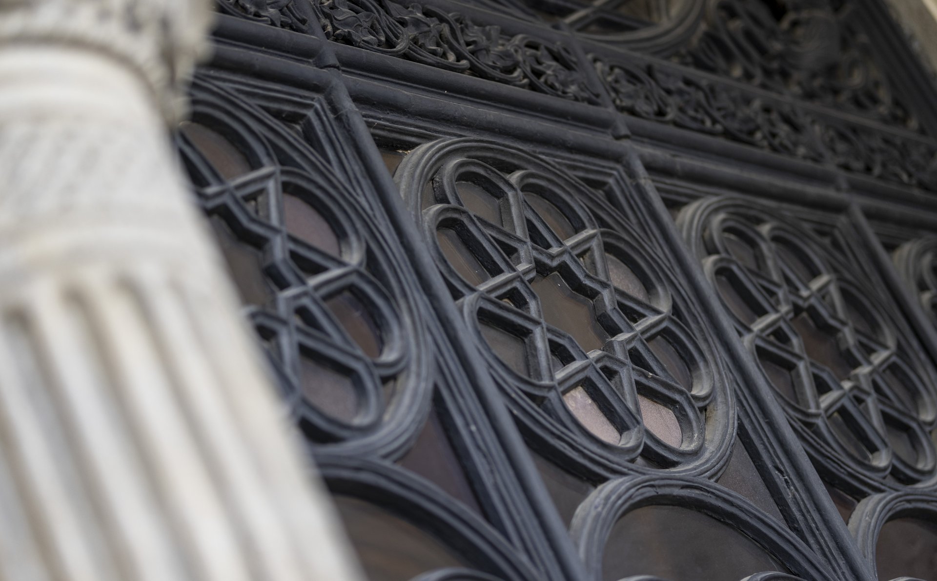 Das Bild zeigt einen Teil der Außenfassade des Heeresgeschichtlichen Museums. Im Fokus steht ein dekoratives Fenster mit kunstvollen, gusseisernen Gittern, die geometrische und florale Muster bilden. Die Fensterrahmen sind dunkel und fein gearbeitet, wobei jedes einzelne Fenster ein einzigartiges Muster zeigt. Im Vordergrund ist ein Teil einer Säule zu sehen, die mit vertikalen Rillen verziert ist. 