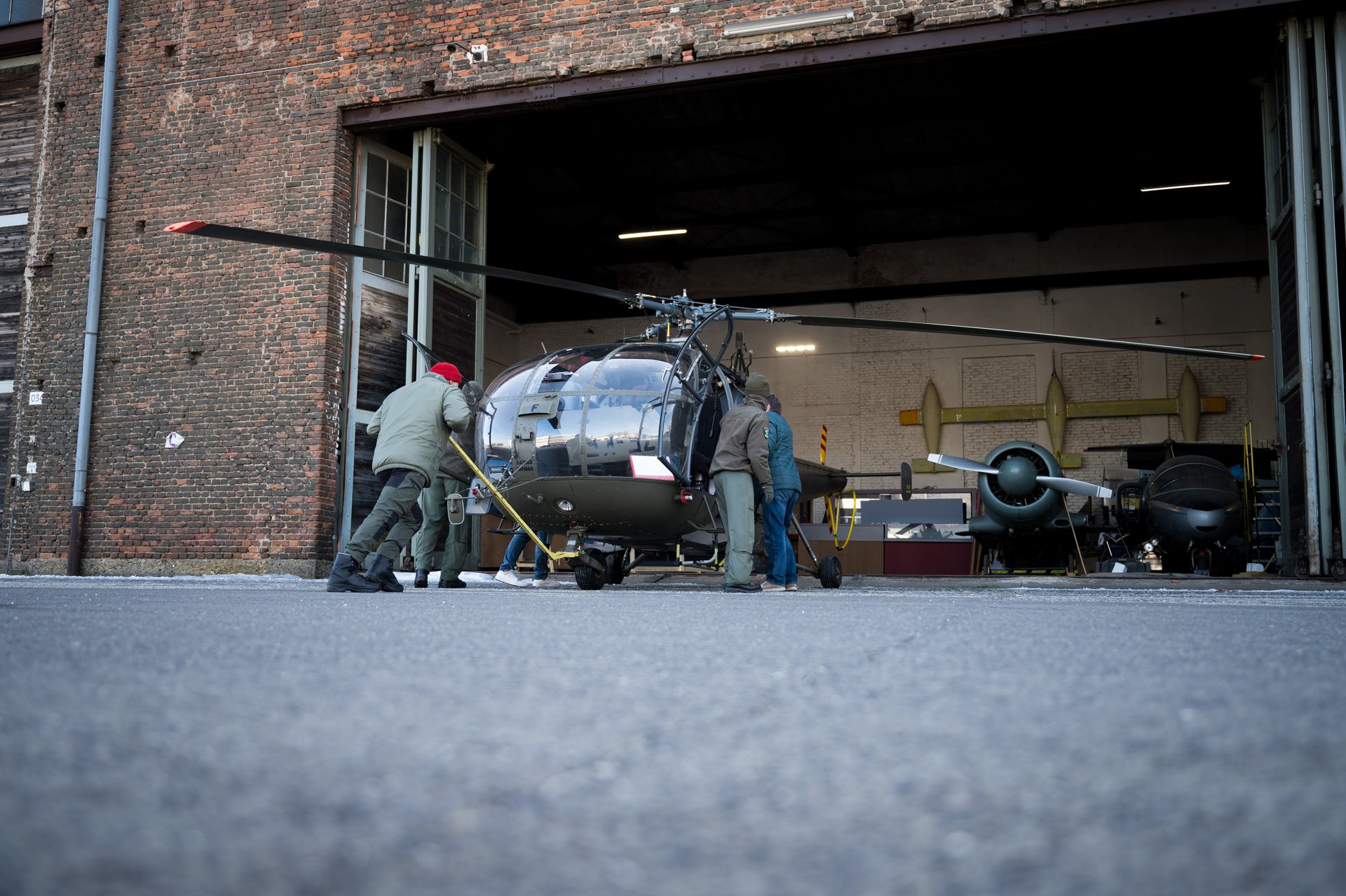 Dieses Bild zeigt das Hangar-Tor in der Ameisenperspektive, durch das ein Militärhubschrauber von mehreren Mitarbeitern geschoben wird. Der Hubschrauber wird langsam und sorgfältig durch den Torbogen geschoben, während die Mitarbeiter ihn unterstützen.