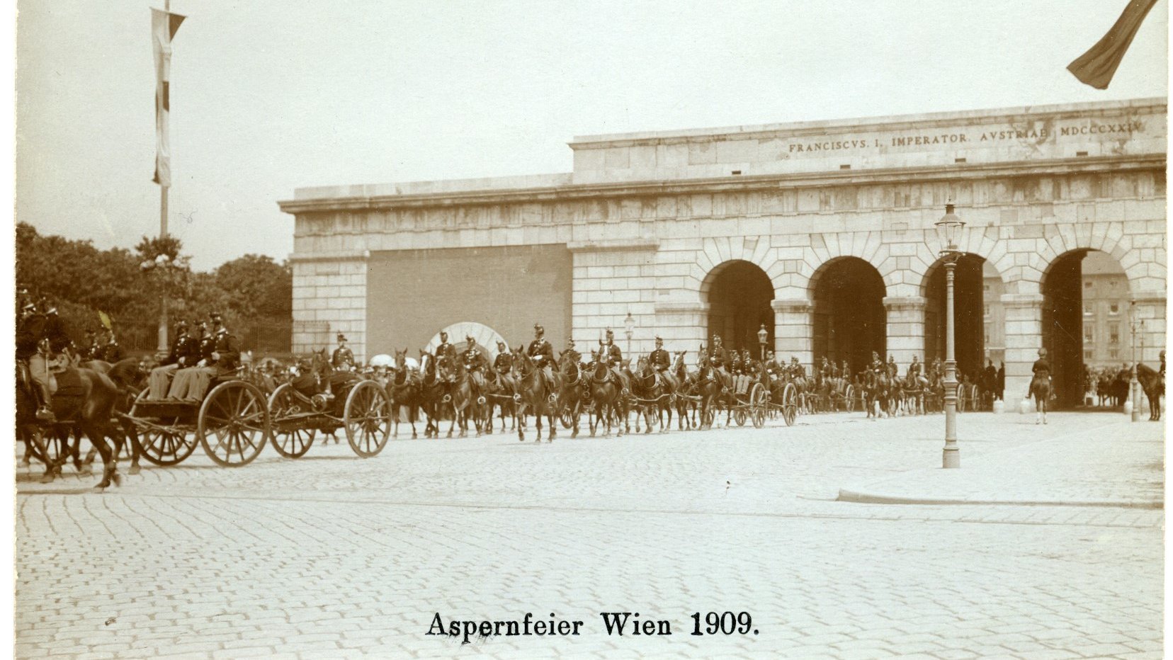 Das Bild zeigt eine militärische Parade, die 1909 anlässlich der Aspernfeier in Wien stattfand. Im Vordergrund marschieren Soldaten und Kutschen mit Pferdegespannen über einen gepflasterten Platz. Im Hintergrund sieht man das Heldentor (Äußeres Burgtor) in Wien, auf dem die Inschrift „Franciscus I. Imperator Austriae MDCCCXXXIII“ zu sehen ist. Die Aspernfeier erinnert an die Schlacht bei Aspern im Jahr 1809, bei der die österreichischen Truppen unter Erzherzog Karl einen Sieg gegen Napoleon errangen.