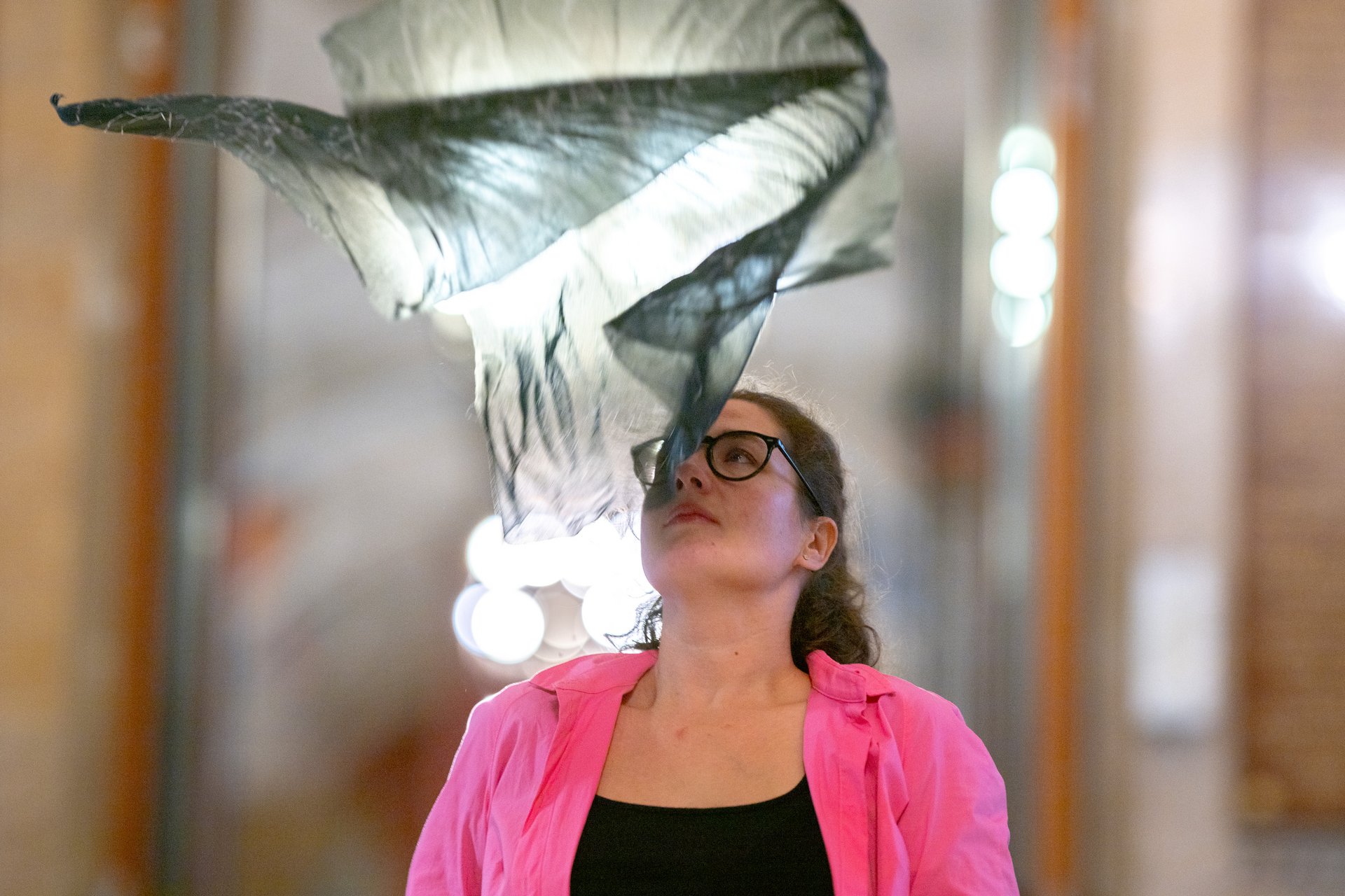 Auf diesem Bild sieht man die Künstlerin Raffaela Bielesch in der Mitte. Über ihren Kopf scheint eine hauchdünne schwarze Decke runterzufallen, als Teil ihrer Performance. Ihr Blick ist nach oben gerichtet. Der Hintergrund ist verschwommen. 