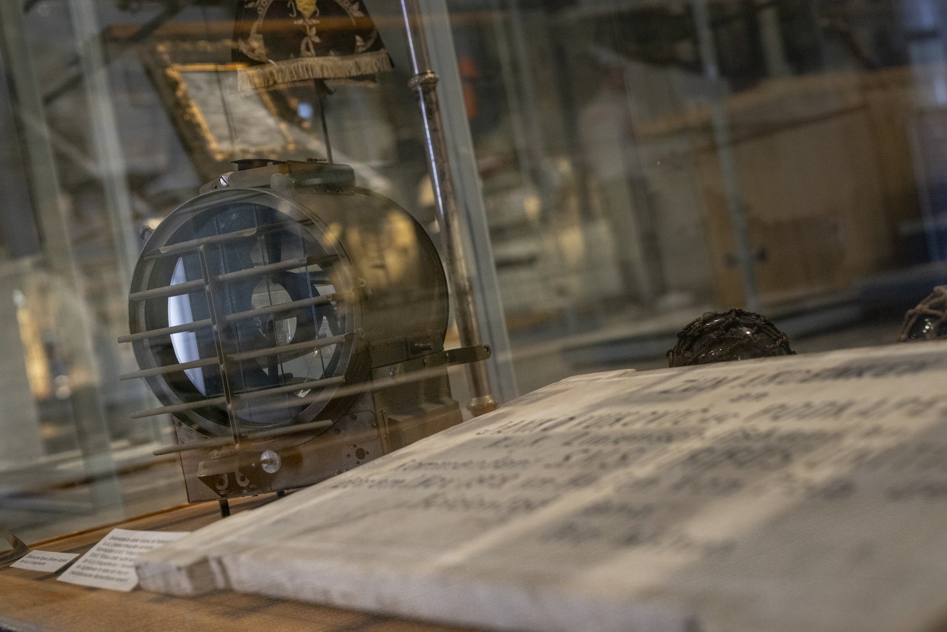 Im Vordergrund ist ein altes maritimes Suchscheinwerfer zu sehen, ein wichtiges Instrument auf historischen Schiffen, das für die Navigation und Kommunikation bei Nacht verwendet wurde. Der Suchscheinwerfer ist in einer Vitrine ausgestellt und von einer Schutzvorrichtung aus Metall umgeben.  Daneben liegt ein aufgeschlagenes, historisches Dokument, das möglicherweise ein Logbuch, eine Seekarte oder ein anderes wichtiges Schriftstück aus der maritimen Geschichte darstellt. Die Schrift darauf ist handgeschrieben und deutet auf eine gewisse Bedeutung des Dokuments hin. 