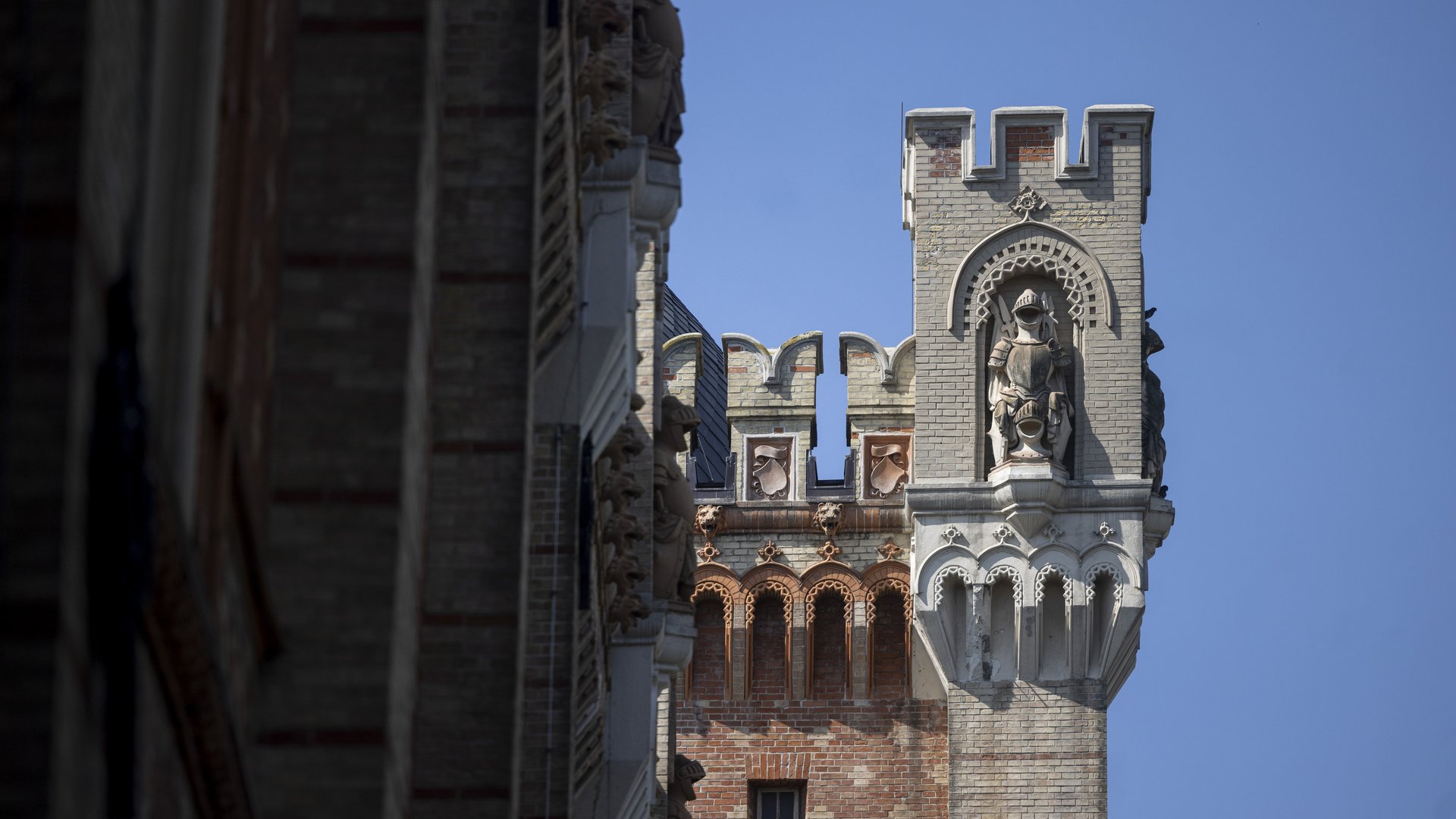 Dieses Bild zeigt ein Teil der Außenfassade des HGMs. Die Fassade zeigt eine Mischung aus Backstein- und Steinelementen. In der Mitte des Bildes befindet sich ein Turm mit einer dekorativen Aussparung, in der eine Ritterstatue steht. Der Ritter trägt ein Schild. Oberhalb der Statue ist ein Bogen mit Mustern zu erkennen. Der Turm hat eine gezackte Zierleiste, die dem mittelalterlichen Design nachgestellt ist. Neben dem Turm sind weitere Gebäudeteile zu sehen, die ebenfalls reich mit Verzierungen und Reliefs ausgestattet sind. Der Himmel ist klar und blau. 