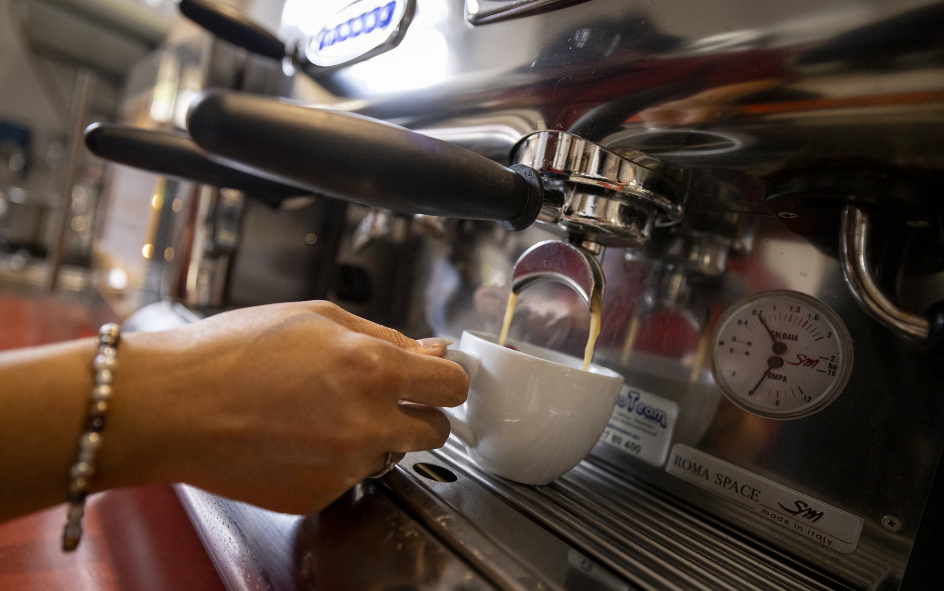 Eine Hand hält eine Kaffeetasse an einem Kaffeevollautomaten. Aus dem Hahn fließt frischer, heißer Kaffee in die Tasse. Die Hand trägt ein Armband und einen Ring am Finger. Der Hintergrund ist verschwommen, der Fokus liegt auf der Kaffeetasse.