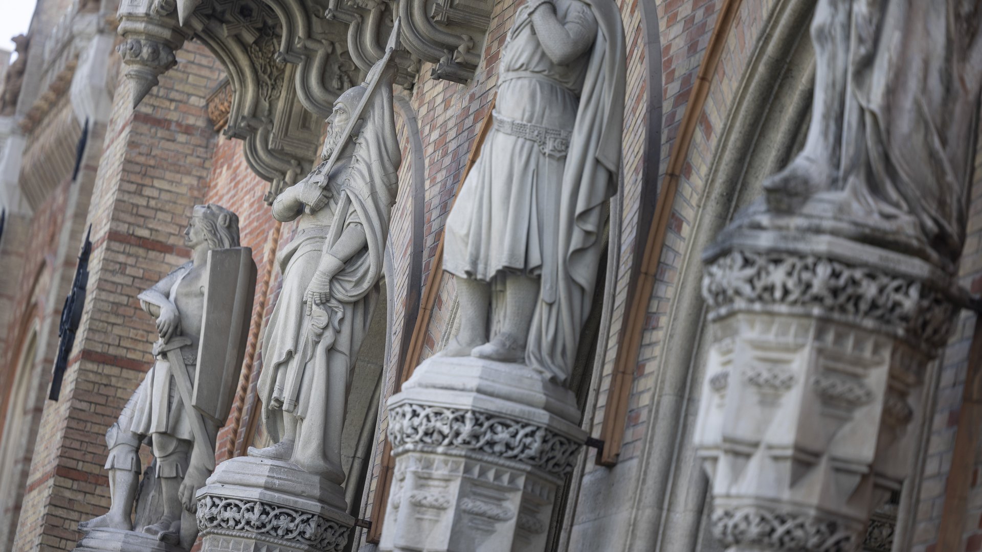 Das Bild zeigt eine Nahaufnahme der Außenfassade des Heeresgeschichtlichen Museums. Zu sehen sind mehrere imposante Steinfiguren, die in regelmäßigen Abständen entlang der Fassade angeordnet sind. Diese Figuren stellen historische Krieger in verschiedenen Posen dar, einige mit Schilden und Schwertern. Sie stehen auf reich verzierten Sockeln, die mit aufwendigen Mustern und Ornamenten versehen sind. Über den Figuren sind dekorative Baldachine angebracht, die ebenfalls mit detaillierten Schnitzereien und architektonischen Verzierungen geschmückt sind. Die Fassade selbst besteht aus roten und beigen Backsteinen, die in einem regelmäßigen Muster angeordnet sind. 