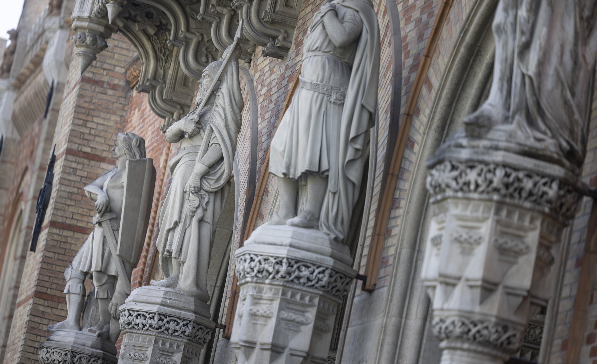 Das Bild zeigt eine Nahaufnahme der Außenfassade des Heeresgeschichtlichen Museums. Zu sehen sind mehrere imposante Steinfiguren, die in regelmäßigen Abständen entlang der Fassade angeordnet sind. Diese Figuren stellen historische Krieger in verschiedenen Posen dar, einige mit Schilden und Schwertern. Sie stehen auf reich verzierten Sockeln, die mit aufwendigen Mustern und Ornamenten versehen sind. Über den Figuren sind dekorative Baldachine angebracht, die ebenfalls mit detaillierten Schnitzereien und architektonischen Verzierungen geschmückt sind. Die Fassade selbst besteht aus roten und beigen Backsteinen, die in einem regelmäßigen Muster angeordnet sind. 