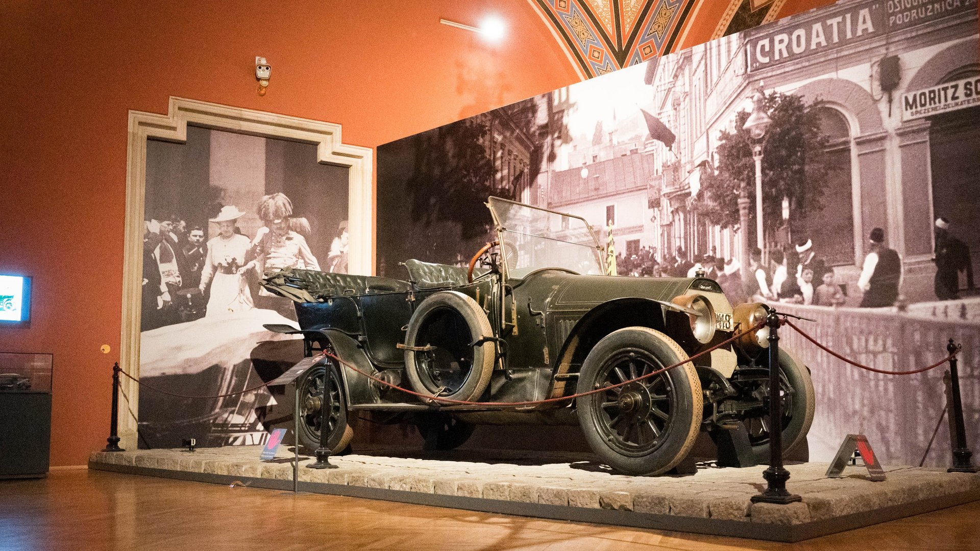 Ein historisches Automobil, ein Gräf & Stift, ausgestellt im Saal "Das Attentat von Sarajevo" des Heeresgeschichtlichen Museums. Das Fahrzeug ist mit einer Absperrung umgeben und auf einem Podest platziert. Im Hintergrund sind großformatige Schwarz-Weiß-Fotografien zu sehen, die Szenen aus der Zeit des Attentats in Sarajevo zeigen. Eine Fotografie zeigt Menschen in historischen Kleidern, eine andere eine Straßenszene mit Gebäuden und dem Schriftzug "CROATIA". Der Ausstellungsraum hat eine warme Beleuchtung und eine verzierte Decke.