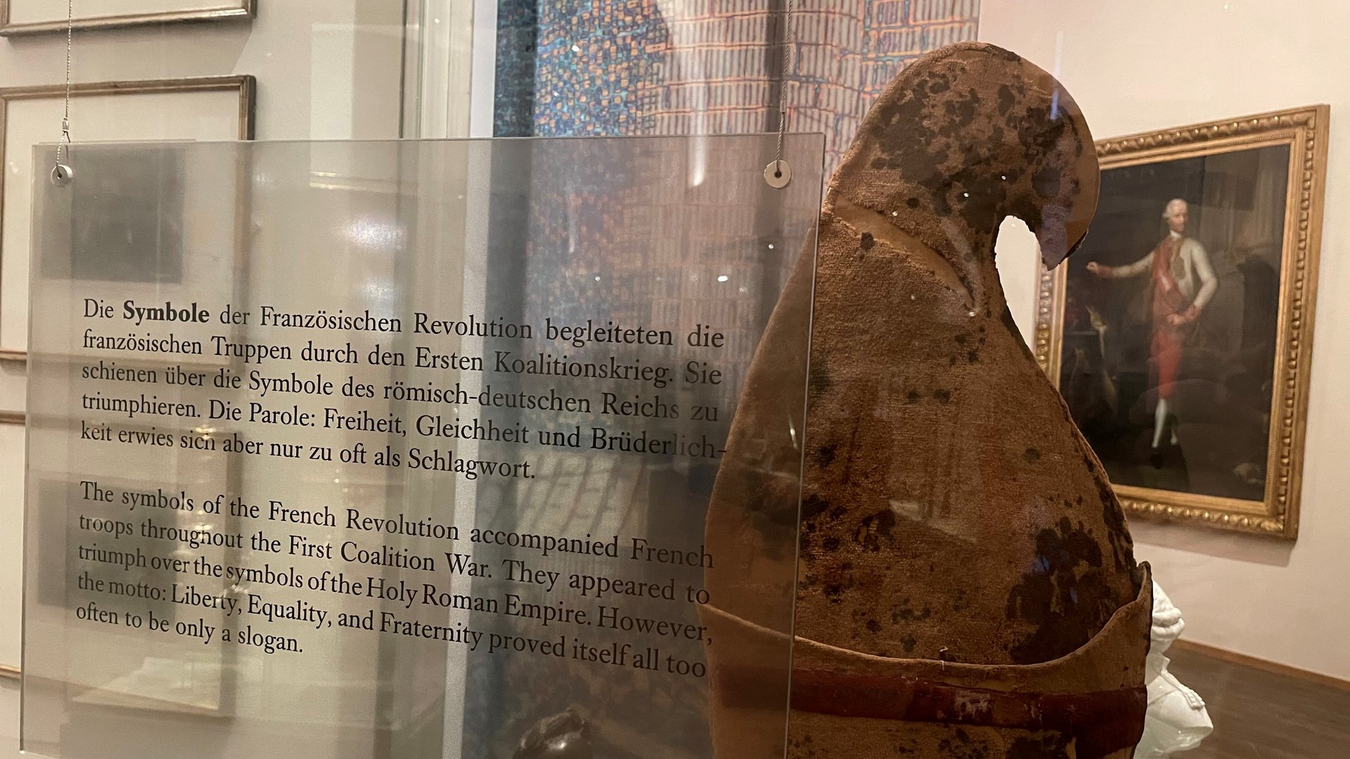 Das Bild zeigt eine Jakobinermütze, die in einer Vitrine ausgestellt ist. Die Jakobinermütze war ein Symbol der Bewegung der Französischen Revolution. Hinter der Vitrine sind gerahmte Lithographien an der Wand zu sehen, die möglicherweise historische Szenen der Revolution darstellen, sowie kleinere Statuetten. 