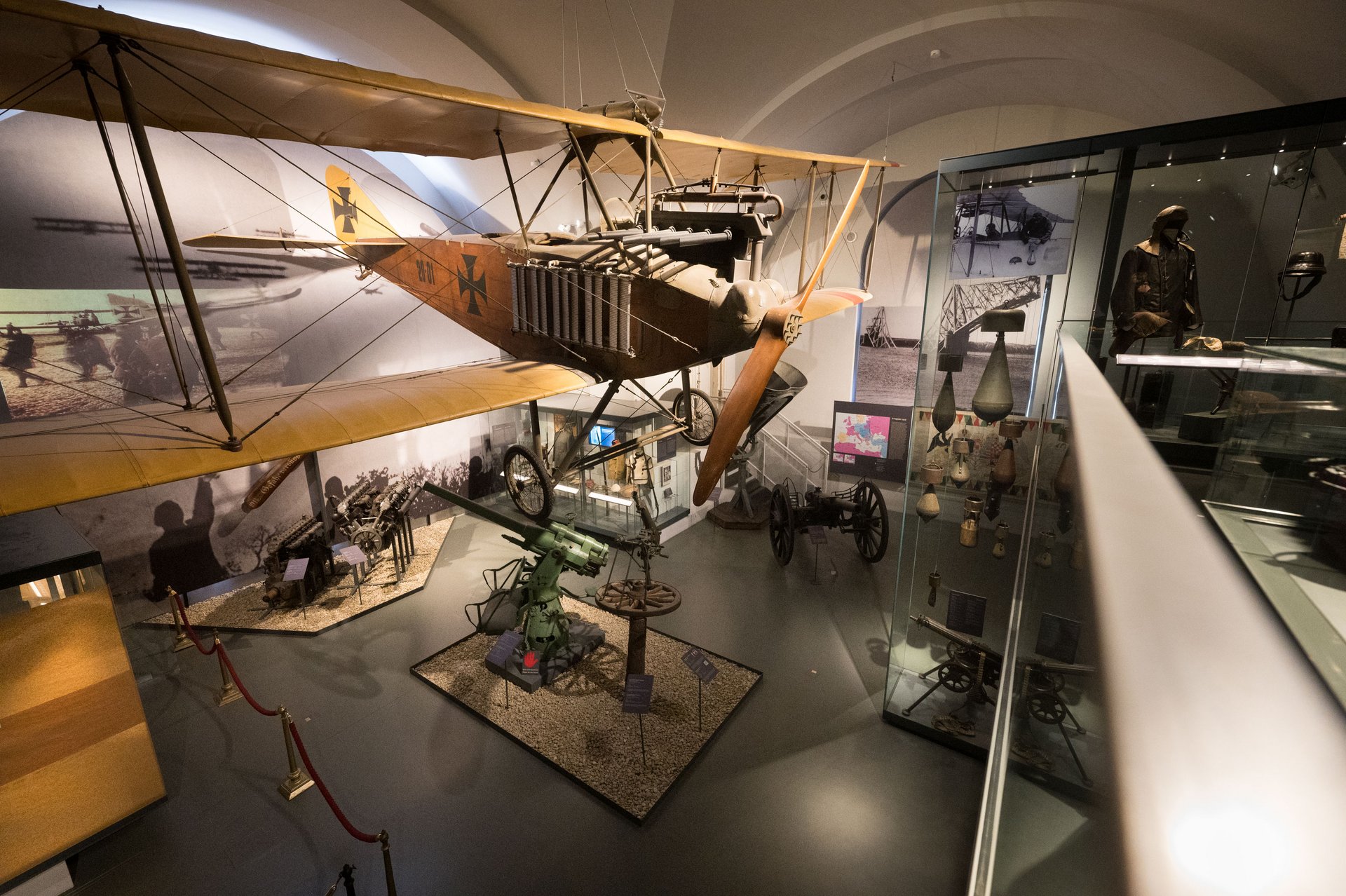 Das Bild zeigt eine Ausstellung im Militärluftfahrtmuseum Zeltweg. Im Zentrum der Aufnahme hängt ein historisches Doppeldeckerflugzeug mit einem Holzkörper und gelben Flügeln von der Decke. Das Flugzeug trägt Markierungen und ein schwarzes Kreuz, das typisch für deutsche Flugzeuge im Ersten Weltkrieg ist. Unter dem Flugzeug sind verschiedene militärische Ausrüstungsgegenstände ausgestellt, darunter Maschinengewehre und Kanonen, die auf Podesten mit Kieselsteinen platziert sind.