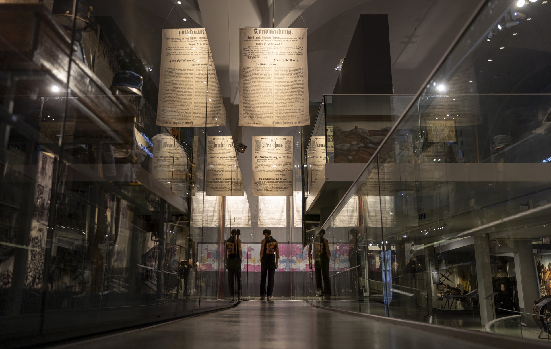 Das Bild zeigt eine Ausstellung im Heeresgeschichtlichen Museum, die sich dem Ersten Weltkrieg widmet. Im Zentrum des Bildes sind große, hängende Zeitungsseiten und Ankündigungen zu sehen, die von der Decke herab hängen und historische Informationen und Nachrichten aus der Kriegszeit darstellen. Der Ausstellungsraum ist modern gestaltet, mit Glasgeländern und reflektierenden Oberflächen, die eine tiefere Dimension und einen interessanten visuellen Effekt erzeugen. Auf beiden Seiten des Ganges sind verschiedene militärische Artefakte und historische Objekte in Vitrinen ausgestellt, darunter Uniformen, Waffen und andere Kriegsgegenstände.  Im Hintergrund sind Besucher zu sehen, die die Ausstellung betrachten.