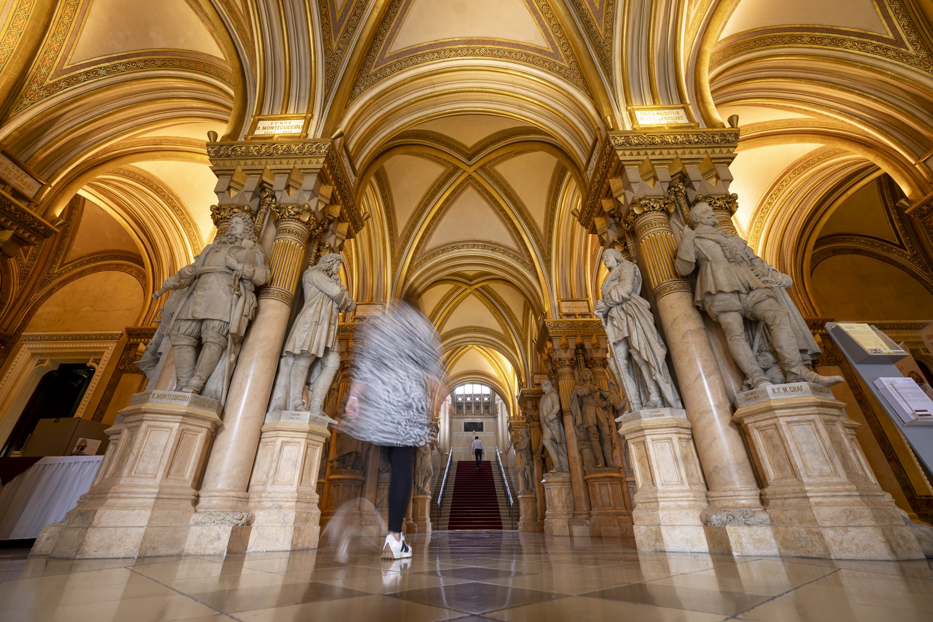 Das Bild zeigt die beeindruckende Feldherrenhalle im Heeresgeschichtlichen Museum. Diese Halle ist bekannt für ihre prächtigen, gewölbten Decken und die Reihe von Statuen bedeutender Militärführer, die entlang der Säulen aufgestellt sind. Die Architektur der Halle ist opulent, mit reich verzierten Bögen und Säulen, die mit goldenen Akzenten und kunstvollen Details geschmückt sind. Die Statuen, die historische Persönlichkeiten und Feldherren darstellen, stehen erhöht auf Sockeln und blicken majestätisch in den Raum. Im Hintergrund führt eine breite Treppe nach oben, was die Erhabenheit und Größe des Raumes zusätzlich betont. Diese Treppe und die symmetrisch angeordneten Statuen schaffen einen eindrucksvollen Anblick, der die Bedeutung und den historischen Wert der Feldherrenhalle unterstreicht.