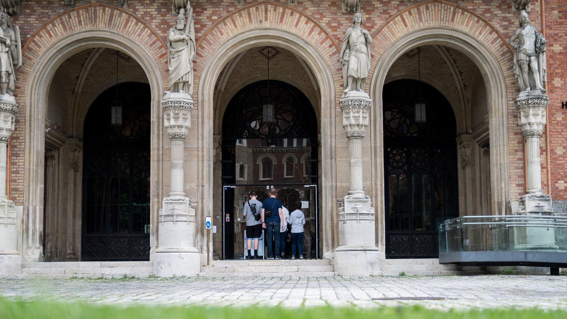 Aus der Ameisenperspektive auf der Wiese ist die Außenfront des Heeresgeschichtlichen Museums mit dem Haupteingang zu sehen. An der Außenfassade des Museumsgebäudes sieht man das Geländer für einen barrierefreien Zutritt für Rollstuhlfahrer. Statuen stehen an den Bogensäulen des Haupteingangs. Der Hof ist mit Pflastersteinen belegt. Vor dem Eingang befinden sich vier Personen in Freizeitkleidung. Ein Paar hält Händchen und betritt gerade durch die Glastür das Gebäude; die Frau trägt eine graue Weste und dunkelblaue Jeans, der Mann trägt einen blauen Pullover und dunkle Jeans. Dahinter gehen zwei weitere Männer: der eine links trägt ein graues Shirt, schwarze Shorts und einen kleinen Rucksack, der andere rechts trägt dunklere Kleidung und einen Turnbeutel.