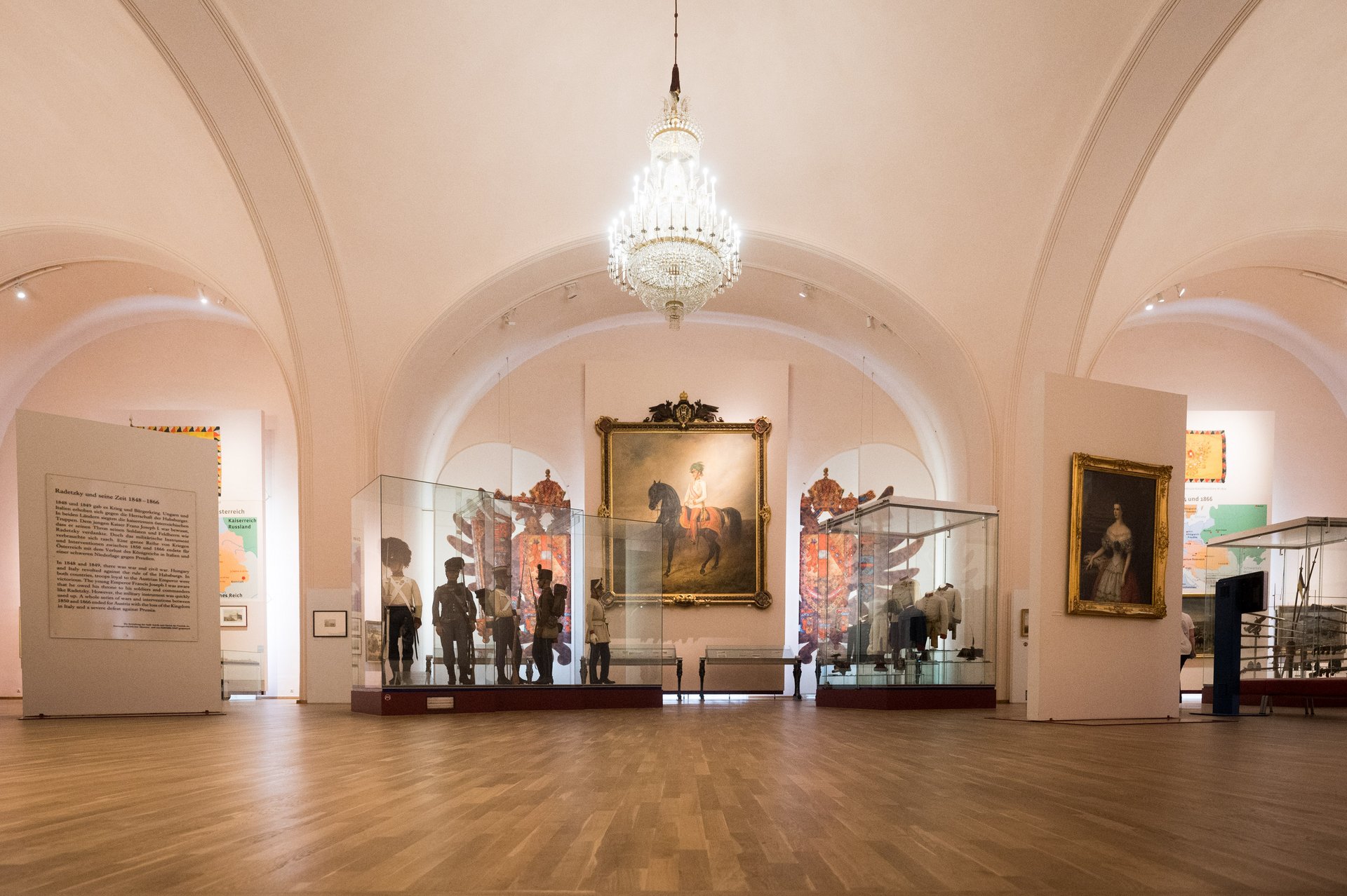 Das Bild zeigt Vitrinen mit Schaufensterpuppen in historischen Uniformen, die die Ära der Revolution von 1848 repräsentieren. Im Hintergrund sind Gemälde, Wappen und Landkarten an den Wänden zu sehen, die die Dauerausstellung über Radetzky illustrieren.