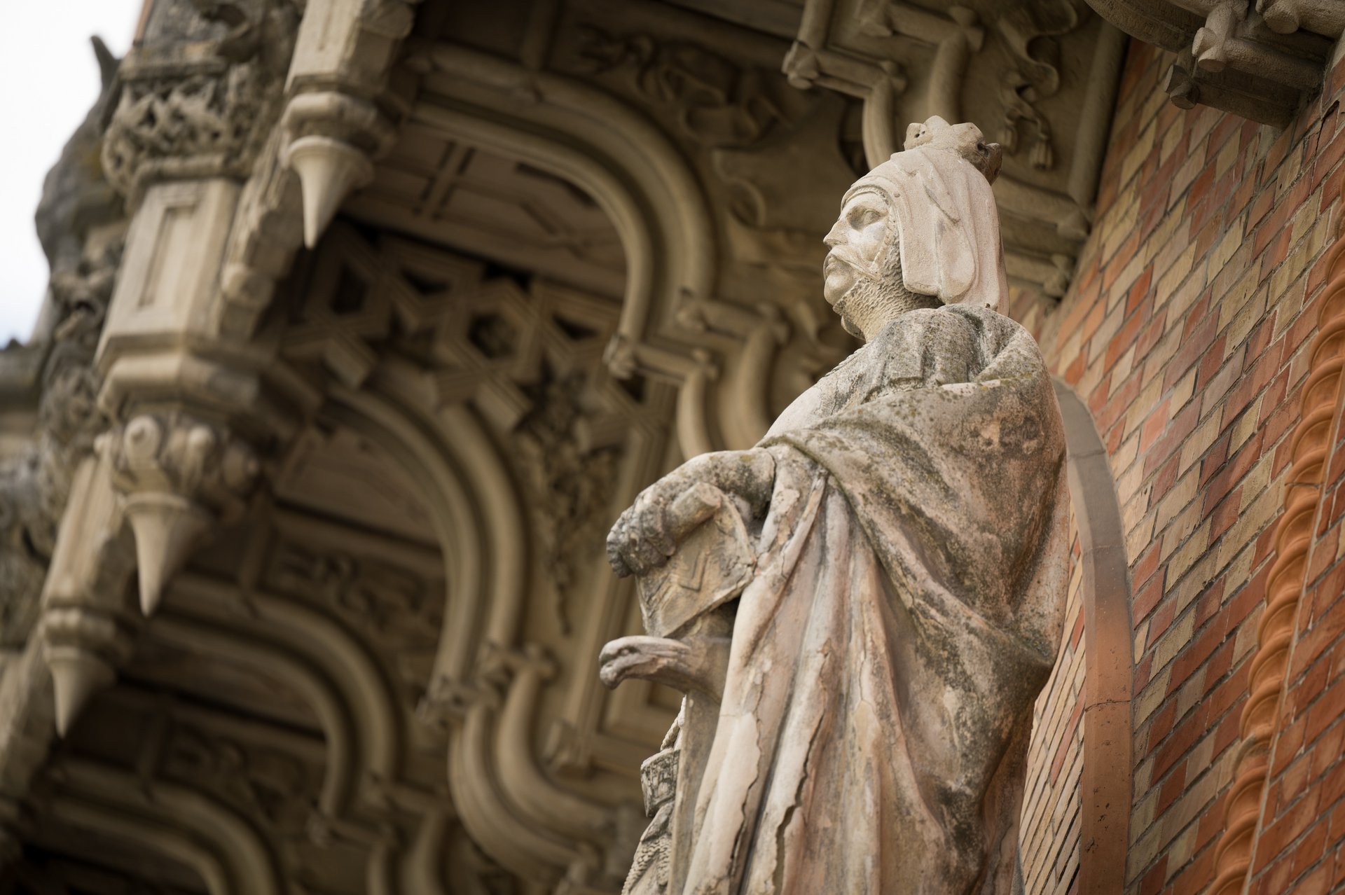 Das Bild zeigt eine detaillierte Nahaufnahme einer beeindruckenden Ritterstatue, die in die Backsteinfassade des HGMs integriert ist. Die links Hand des Ritters ruht auf einem Schwert, während die andere Hand auf einem Dolch ruht, der teilweise versteckt ist. Die Statue steht auf einem floral gestalteten Sockel. Die umgebene Backsteinmauer ist in einem warmen Rotton gehalten und zeigt ein regelmäßiges Muster von helleren und dunkleren Steinen. Ein horizontal verlaufendes Band verläuft über die gesamte Breite der Fassade. 