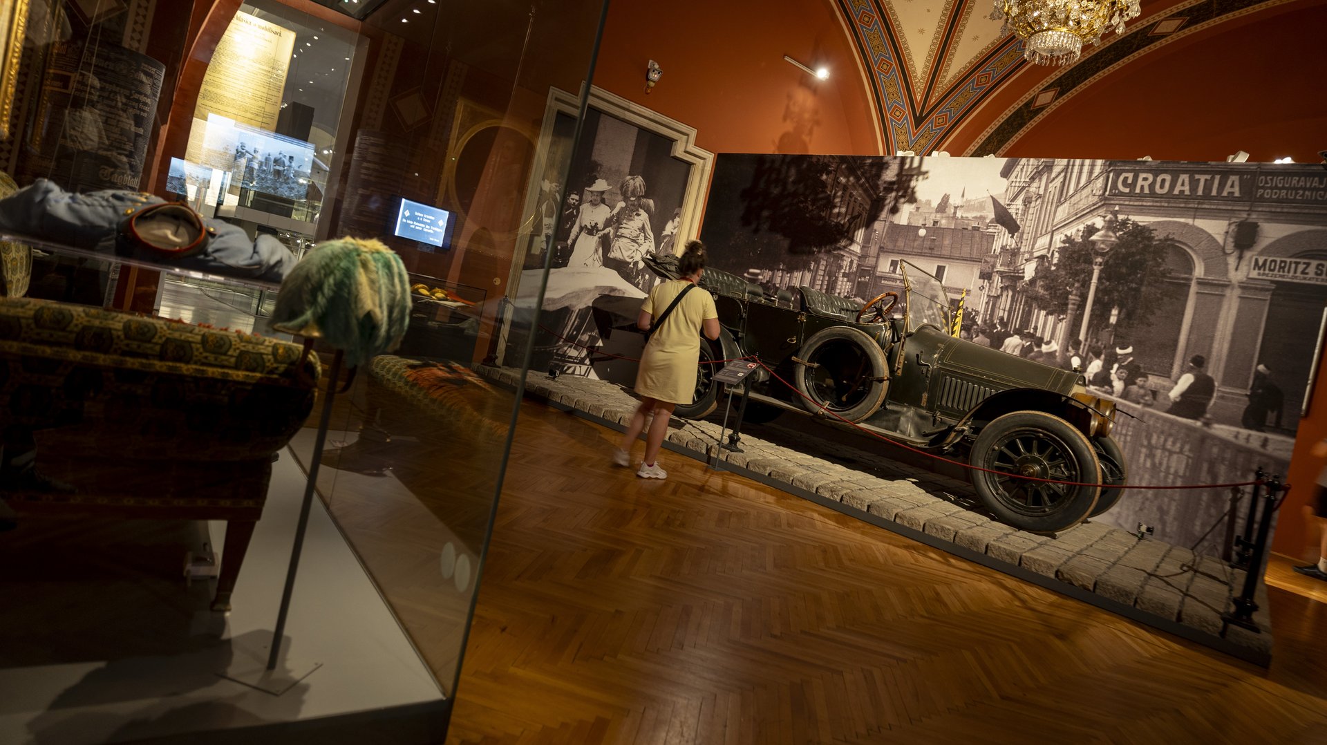 Das Bild zeigt das Gräf & Stift Automobil, in dem der Thronfolger Erzherzog Franz Ferdinand und seine Frau Sophie Chotek am 28. Juni 1914 ermordet wurden, aus der Ameisenperspektive betrachtet. Die Szene ist von einem passend roten Anstrich an den Wänden und dem hölzernen Fußboden umgeben, während die Beleuchtung die Ausstellungsstücke eindrucksvoll hervorhebt. Links befinden sich die Uniform und die Chaiselongue, auf der der Thronfolger seinen schweren Verletzungen erlag. Diese Ausstellungsstücke bieten einen eindrucksvollen Einblick in die tragischen Ereignisse von Sarajevo, die den Ausbruch des Ersten Weltkriegs auslösten und die Geschichte Europas maßgeblich prägten.