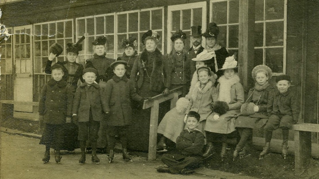 Das Bild zeigt eine Gruppe von Kindern und Erwachsenen, die in warmer Winterkleidung vor einem Holzgebäude mit großen Fenstern posieren. Die meisten tragen Mäntel, Hüte und Muffs, passend zur kalten Jahreszeit. Es scheint eine Winteraktivität oder ein Treffen im Freien zu sein. Die handschriftliche Notiz auf der rechten Seite des Bildes lautet: „Erinnerung an den Winter 1900 - 1901 im Arsenal, Eis. Club.“, was darauf hindeutet, dass es sich um eine Erinnerung an ein Ereignis aus dieser Zeit handelt, vermutlich in einem Eislaufclub.