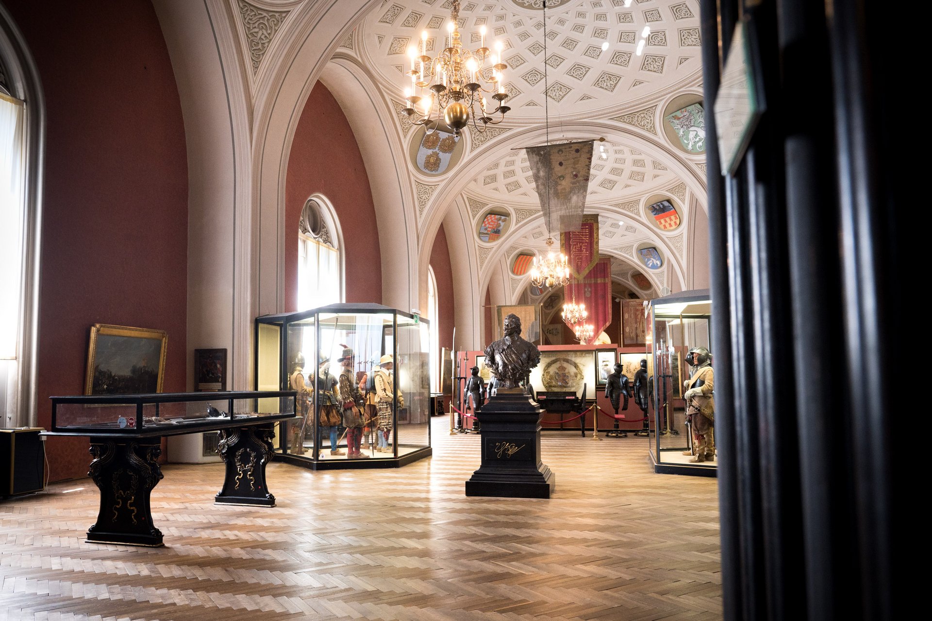 Dieses Bild zeigt eine Ausstellung mit einer braunfarbenen Statue von Prinz Eugen in der Mitte. Dahinter stehen auf jeder Seite jeweils zwei dunkelfarbene Ritterrüstungen. Neben den beiden Rüstungen befinden sich jeweils eine Vitrine mit Puppen in mittelalterlicher Kleidung in gegenüberstehender Stellung. Über der Statue hängt ein Kronleuchter, der den Raum beleuchtet. Der Boden ist hölzern und die Wände sind rot bemalt. Im Hintergrund hängen prachtvolle Wappen an der Wand.