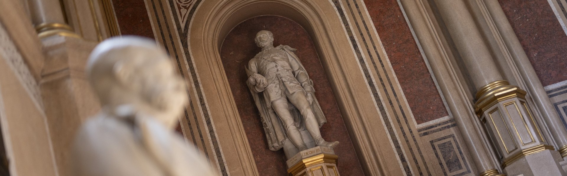 Das Foto zeigt einen Ausschnitt der Feldherrenhalle im Heeresgeschichtlichen Museum (HGM) in Wien. Im Vordergrund befindet sich eine weiße Marmorbüste von Kaiser Franz Joseph, die unscharf abgebildet ist. Im Hintergrund ist eine große Statue eines Feldherren zu sehen, die in eine Nische eingebettet ist. Diese Statue steht auf einem reich verzierten Podest mit goldenen Verzierungen. Die Wand hinter der Statue ist aufwendig gestaltet, mit dekorativen Elementen und Marmorpaneelen, die von Säulen flankiert werden.