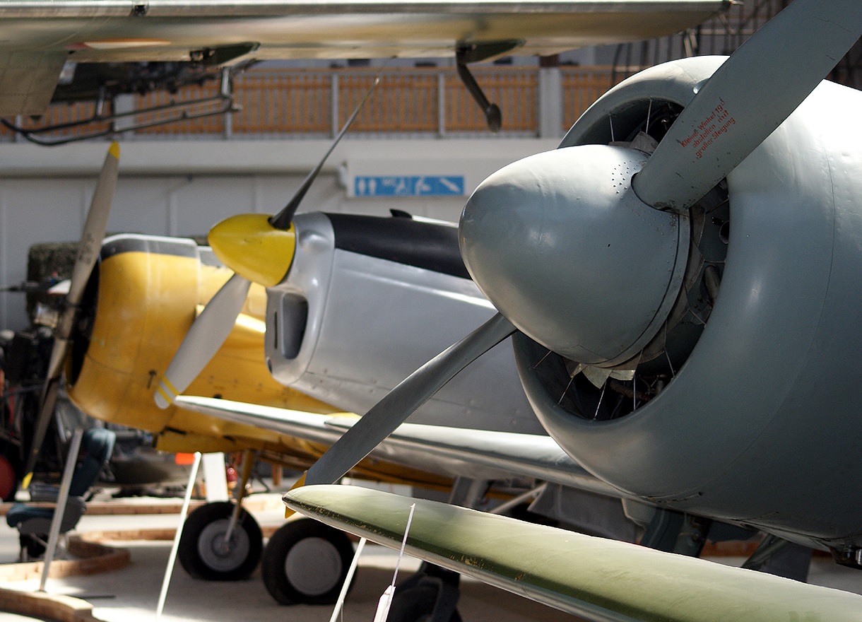 Dieses Bild zeigt drei Propellermaschinen nebeneinander ausgestellt im Luftfahrtmuseum. Die vordere Maschine hat eine graue Lackierung, die mittlere eine Mischung aus grau, schwarz und gelben Farben, während die hintere komplett gelb lackiert ist.